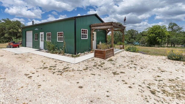 A green building with a garage, a wooden pergola, and a gravel driveway. - The Stargazer Cabin - A Hill Country Retreat (Fredericksburg)