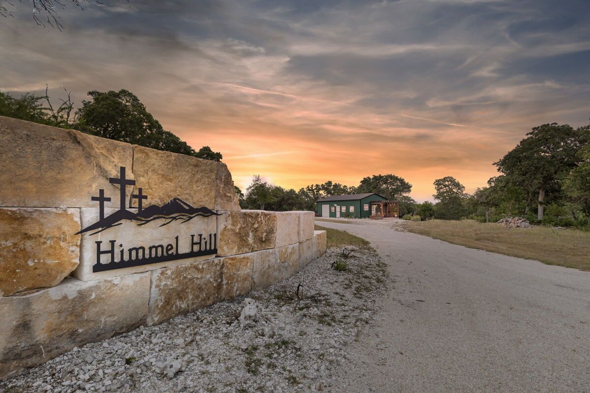 A sign for 'Himmel Hill' with a cross and mountain design, a gravel path, and a building in the background.