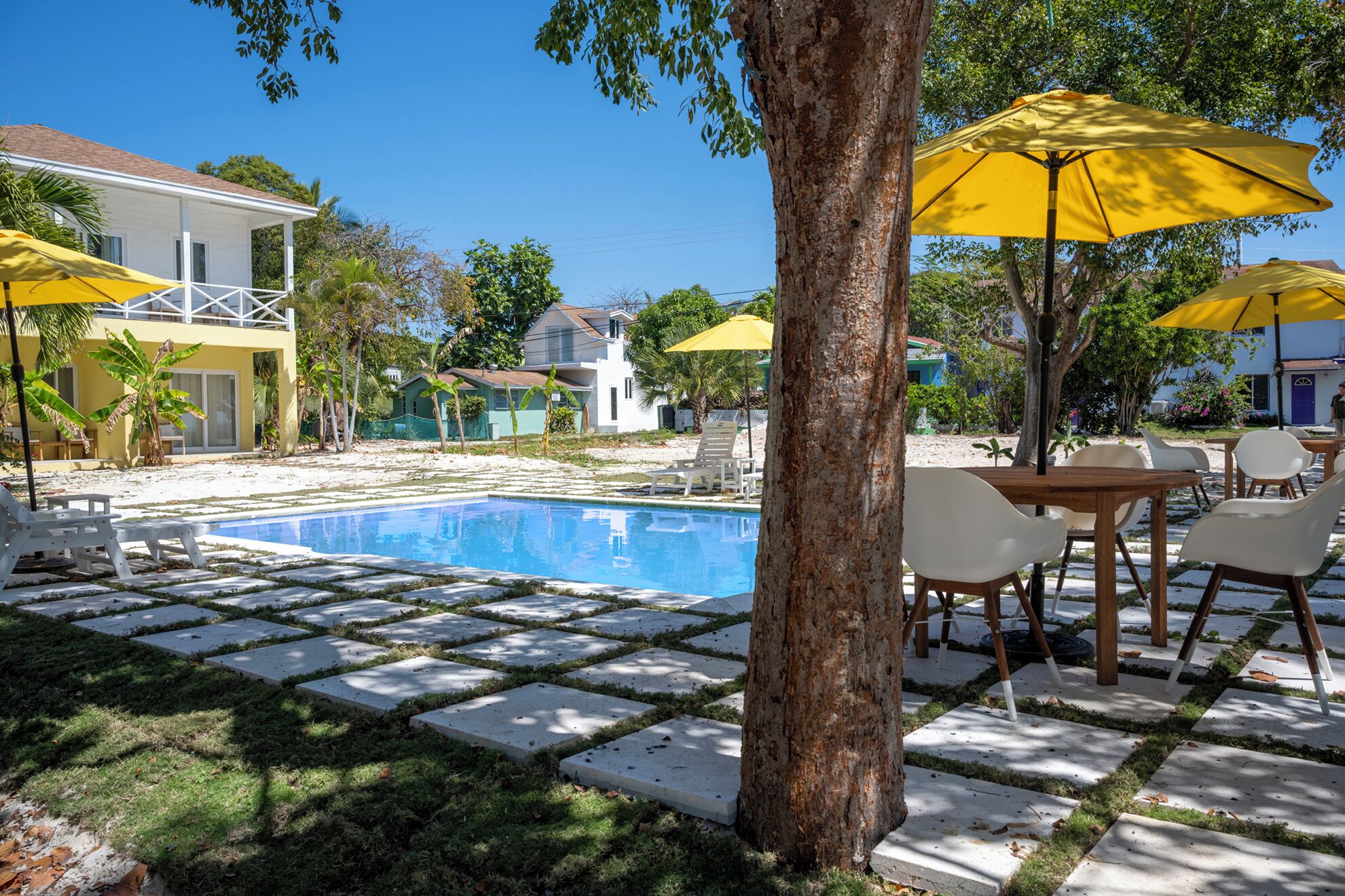 A swimming pool with a tiled deck, surrounded by white lounge chairs and yellow umbrellas, with buildings in the background.