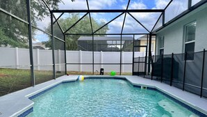 Una piscina con zona techada, rodeada por una valla blanca y una casa al fondo.