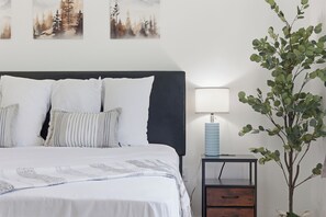 Ein Schlafzimmer mit einem Bett, Kissen, einem Nachttisch mit Lampe und einer Wandpflanze.