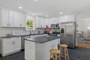 A modern kitchen with white cabinetry, a central island, stainless steel appliances, and a view of greenery outside. - NEW HOUSE • Hot Tub • Game Room • Fire Pit • Sleeps 12 • Near Lakes & Pools (Long Pond)