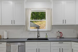 Eine Küche mit weißen Schränken, einer Edelstahl-Spüle, einem Fenster mit Blick auf Bäume und einem Wasserkocher auf der Arbeitsfläche.