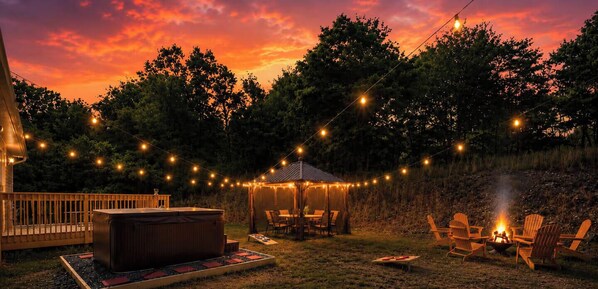 Un área de patio exterior con un jacuzzi, luces colgantes y un hogar para fogata.