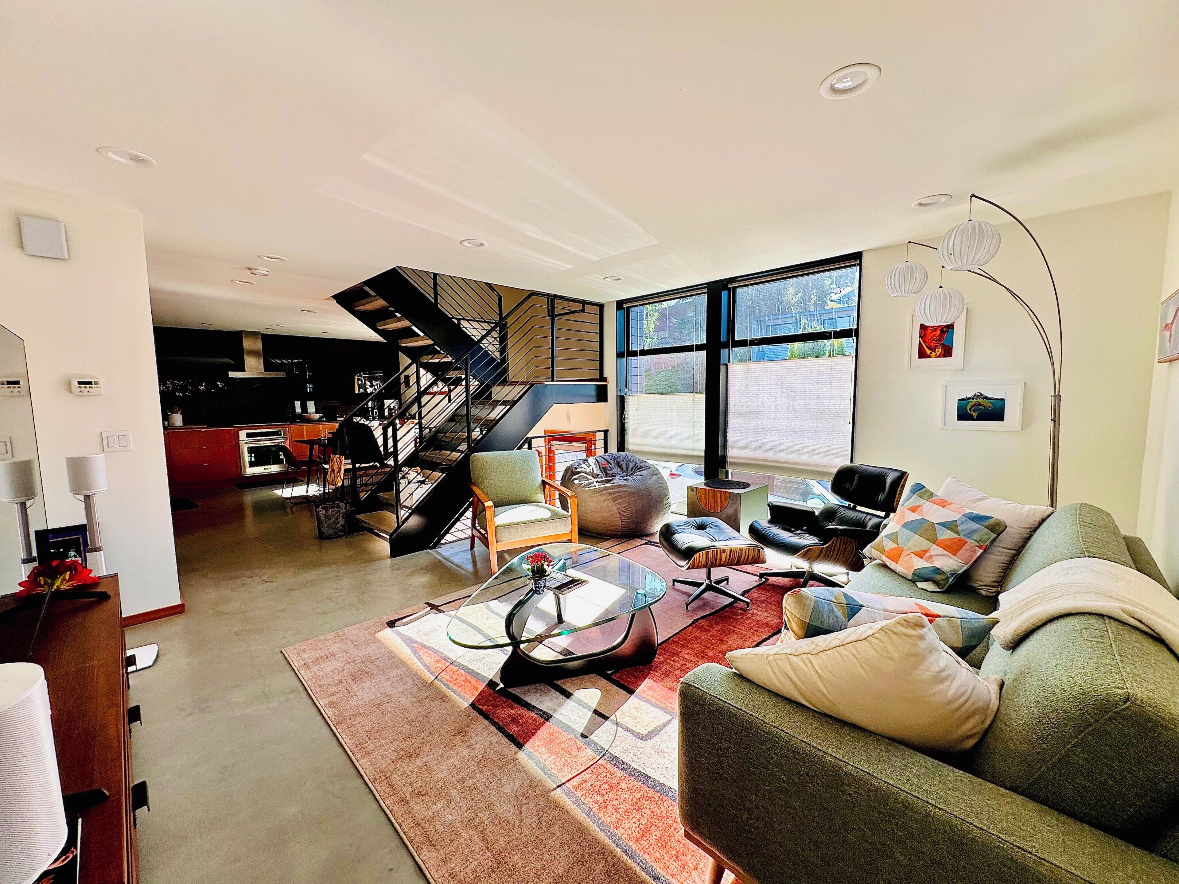 A modern living room with a staircase, a dining area, and large windows.
