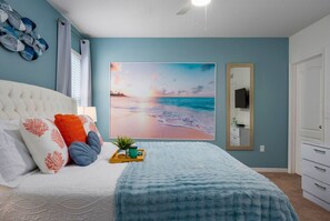Une chambre à coucher avec un grand tableau sur le thème de la plage, une tête de lit blanche à motifs et une commode blanche.