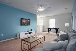 A living room with a blue wall, a white entertainment unit, a glass coffee table, a grey sofa, a pool table, and a ceiling fan. - PROMO read below-Near Disney -Private POOL (Davenport)
