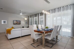 Un espace repas moderne avec une table blanche dressée pour quatre personnes, un canapé agrémenté d’un plaid jaune et un ventilateur de plafond.