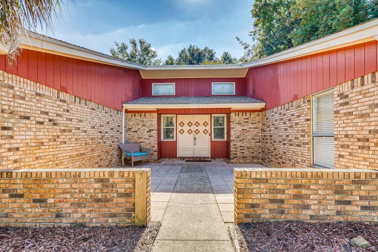 Une maison en briques avec une façade rouge, une porte d’entrée ornée de motifs en losange et un petit patio avec une chaise.
