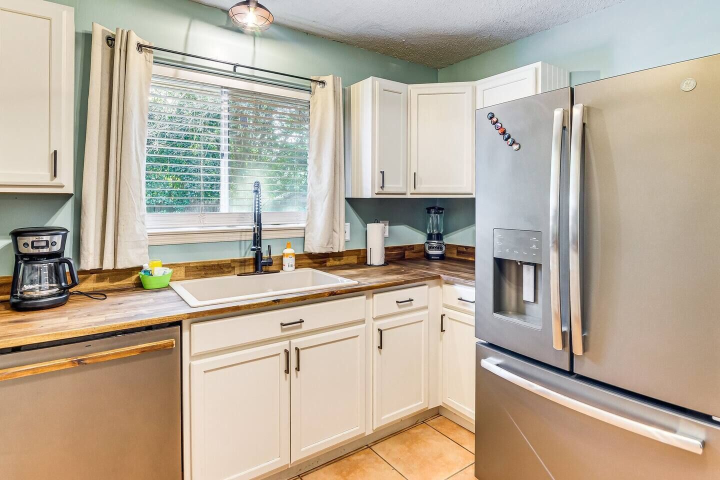 Una cocina con armarios blancos, un refrigerador de acero inoxidable, una cafetera y una ventana con persianas.