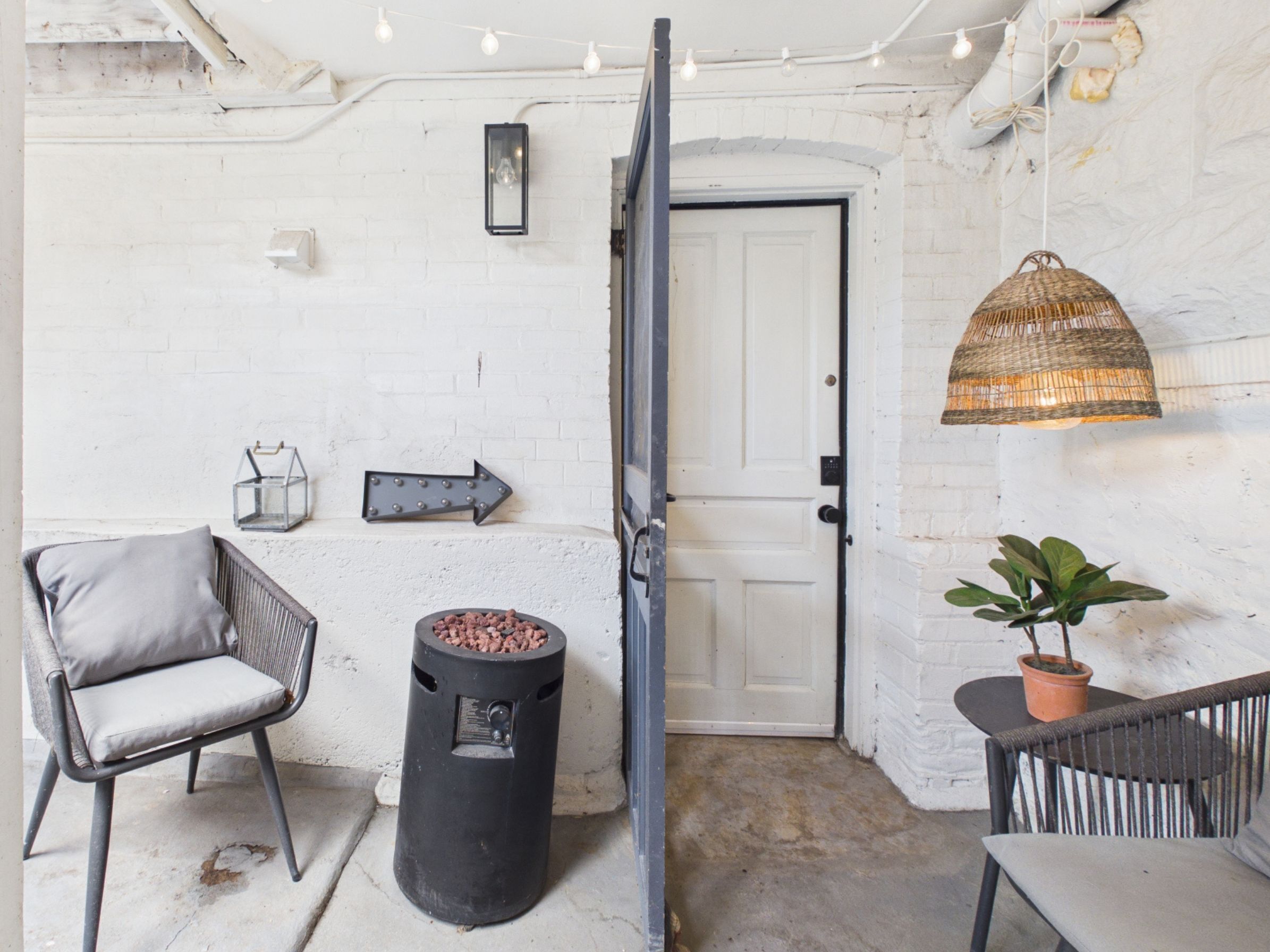 A room with a white brick wall, a door, a black trash bin, a wicker chair, a potted plant, and a hanging light fixture.