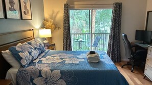 A hotel room with a bed, a desk, a chair, a lamp, and a balcony with a view of trees. - Sunshine Vacation Retreat: Spacious 3-bedroom condo in serene Kissimmee with AC (Kissimmee)