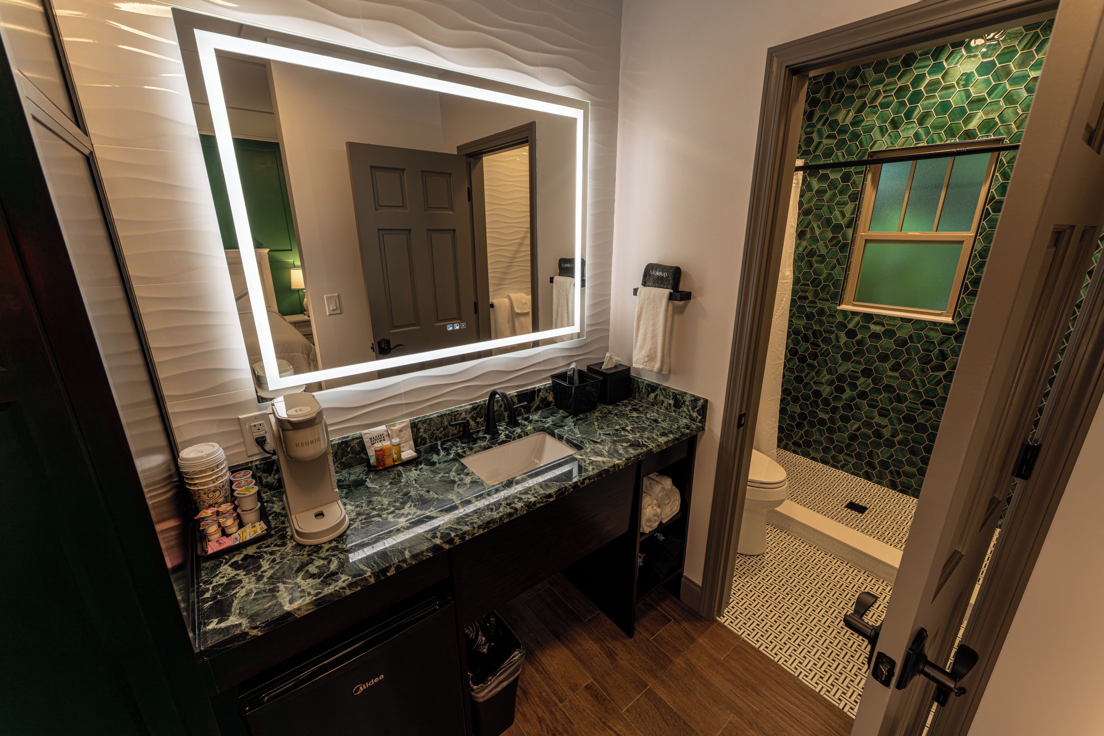 Un baño con encimera de mármol, lavabo, espejo con iluminación, toallero y zona de ducha con pared de azulejos verdes.