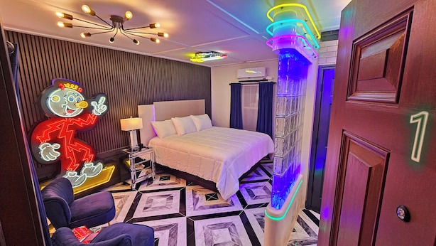Een hotelkamer met een bed, een nachtkastje, een lamp, een neonbord en een deur met een nummer.