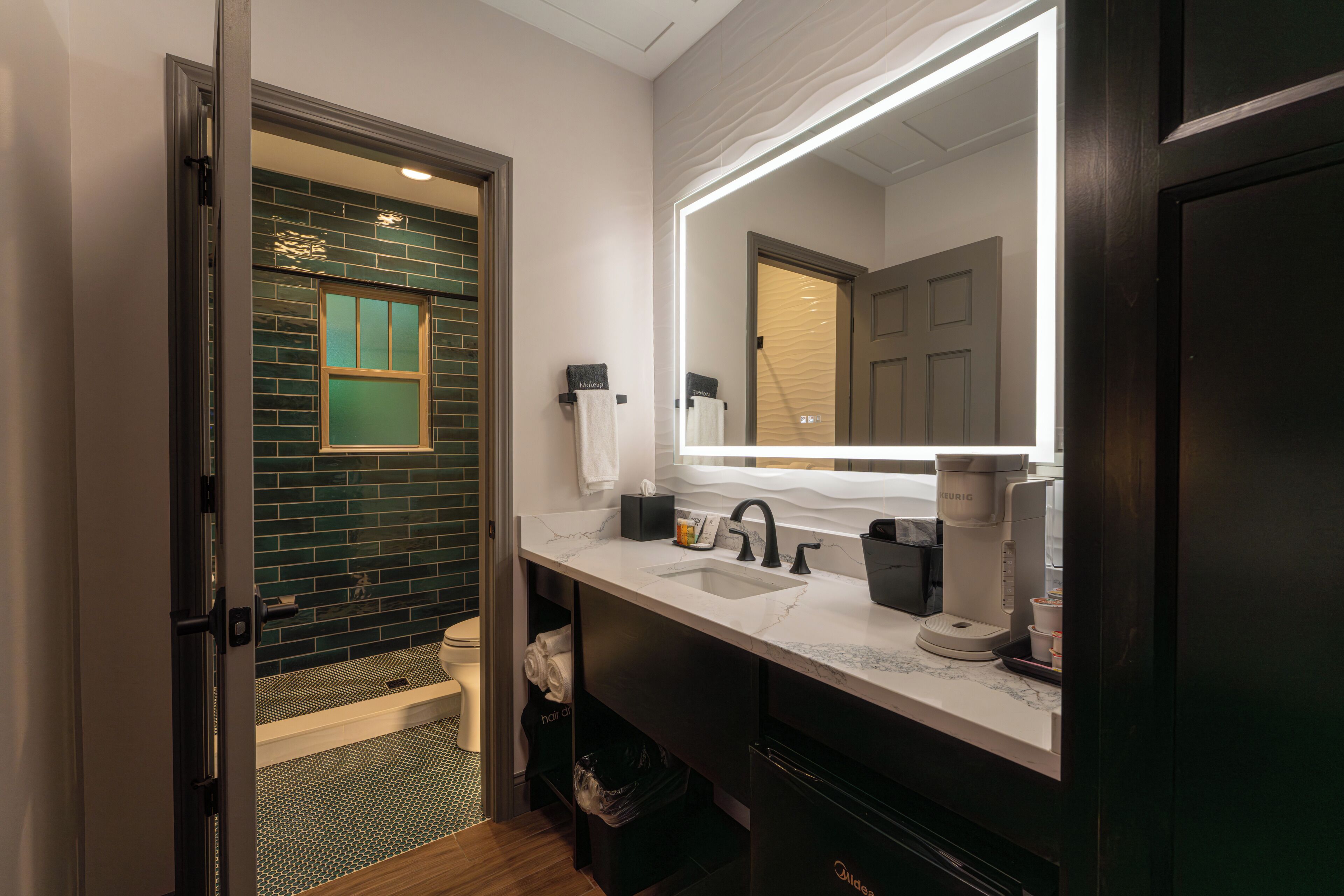 Un baño moderno con un espejo grande, lavamanos, cafetera y zona de ducha.