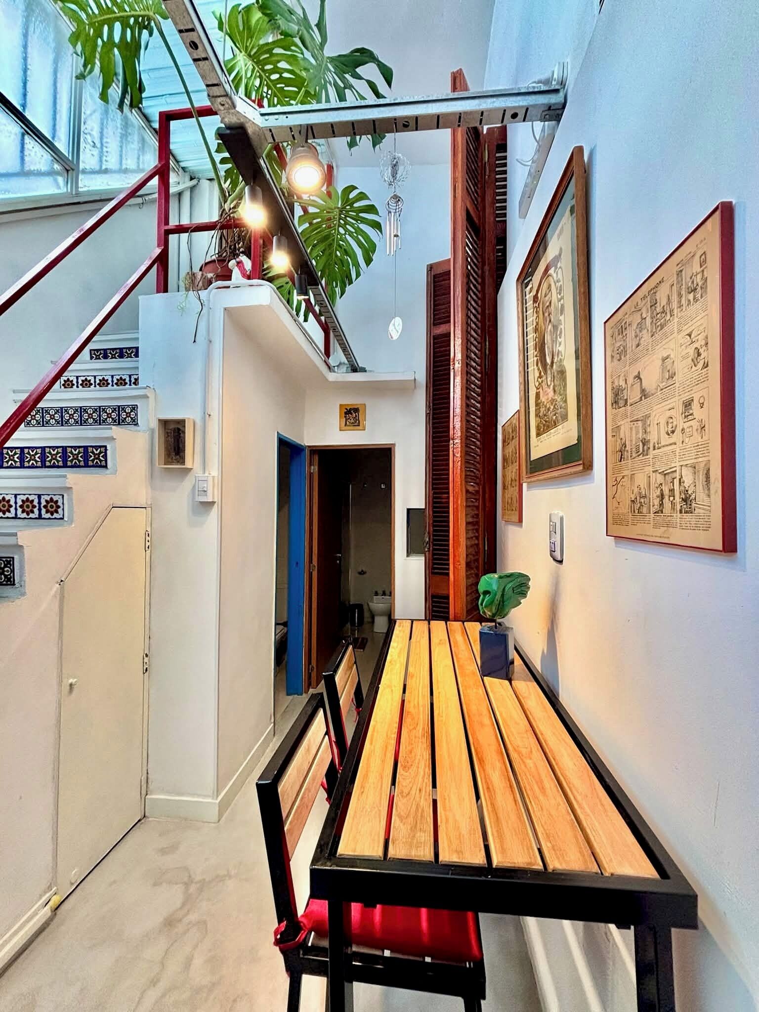 A narrow hallway with a wooden bench and table, a staircase with red railings, and a ceiling with hanging plants and lights.