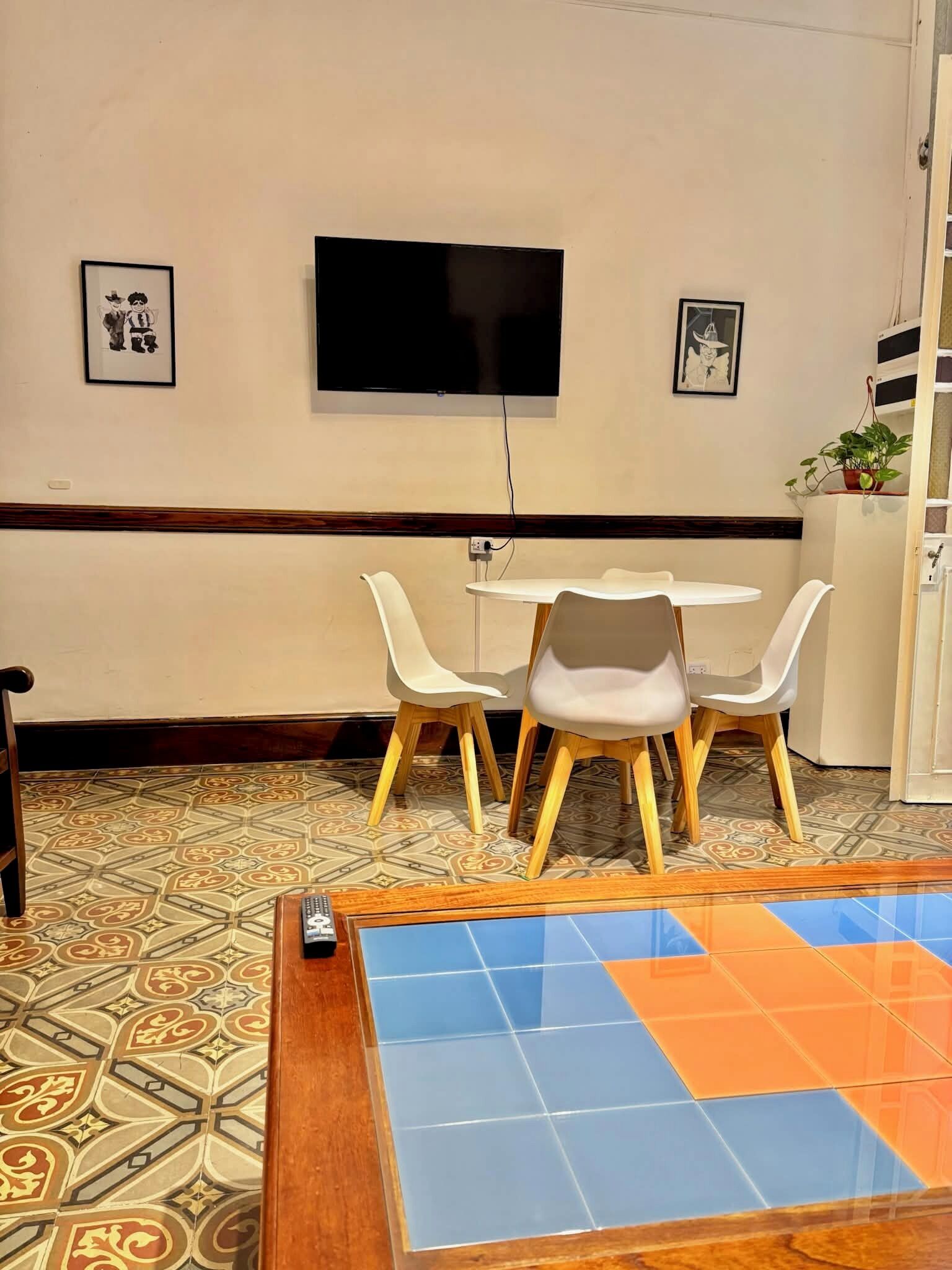 Una habitación con un televisor montado en la pared, una mesita con dos sillas y un suelo estampado.