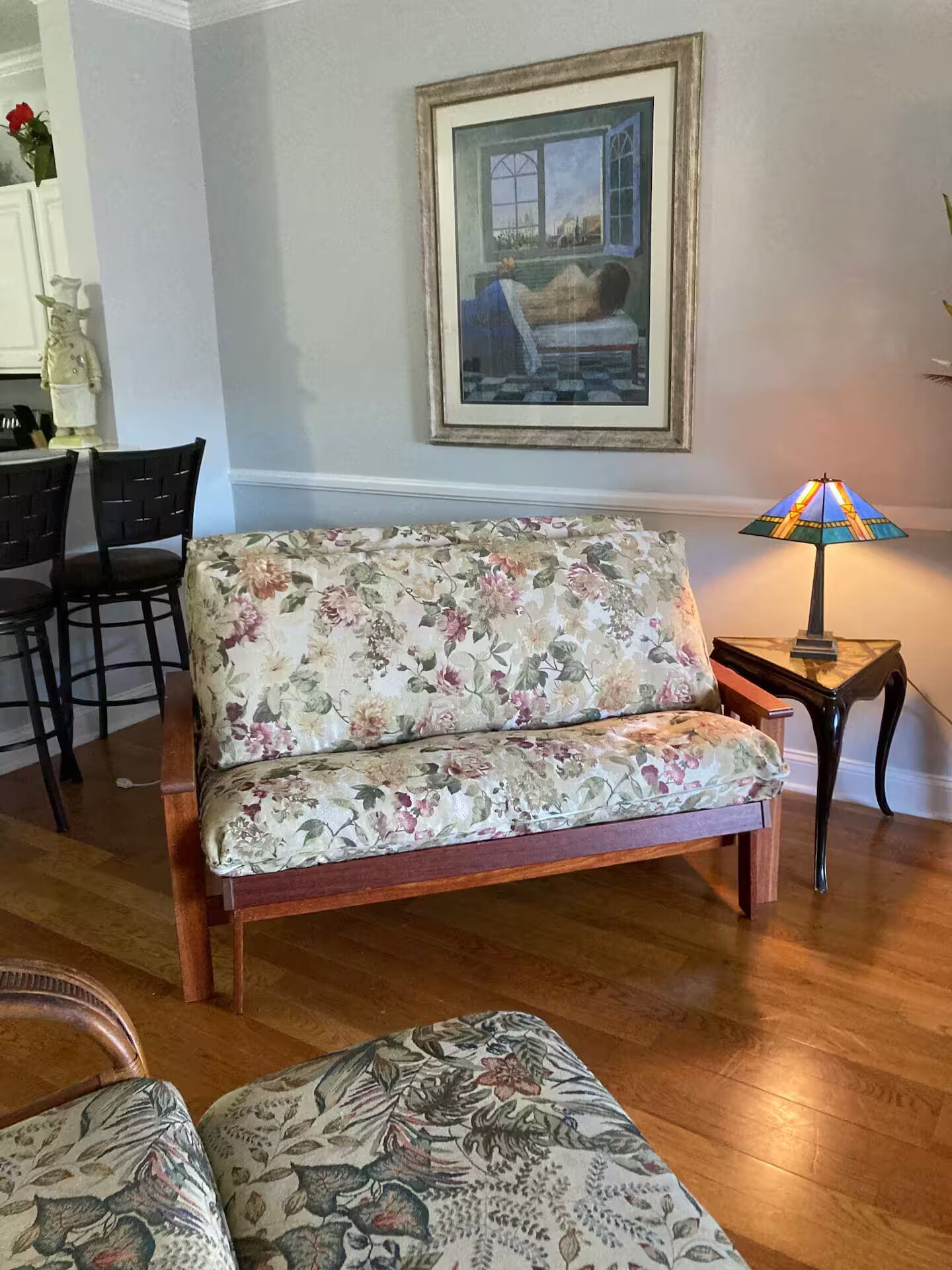 Una sala de estar con un sofá floral, un cuadro enmarcado, una lámpara y un comedor con sillas.