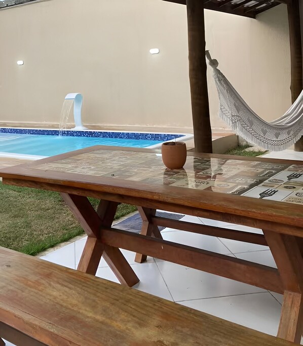 A wooden table with a newspaper, a hammock, and a small pot on a patio with a pool in the background.