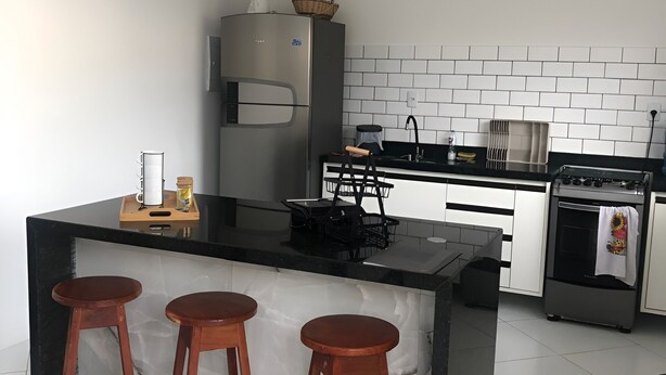 A modern kitchen with a central island, bar stools, a refrigerator, a stove, and a microwave. - Beach house in Jacuípe | Bahia coast | 4 bedrooms | Pool and gourmet area (Camaçari)
