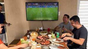 A group of people seated around a wooden table with food and drinks, watching a soccer game on a mounted TV. - Beach house in Jacuípe | Bahia coast | 4 bedrooms | Pool and gourmet area (Camaçari)
