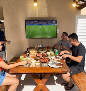 Um grupo de pessoas sentadas em torno de uma mesa de madeira com comida e bebidas, assistindo a um jogo de futebol na televisão instalada.