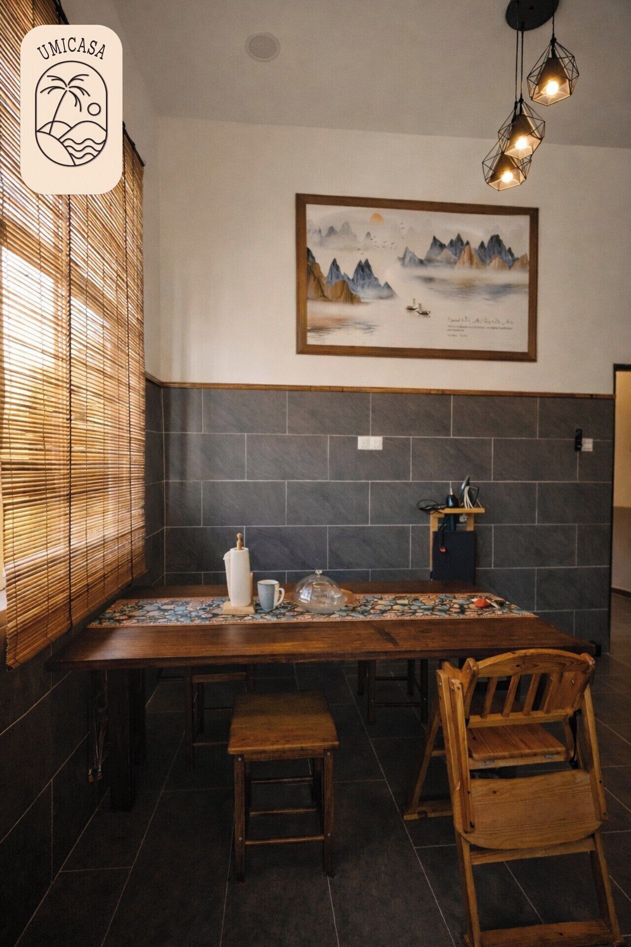 Un coin repas comprenant une table en bois, deux chaises en bois, un store en bambou, un tableau encadré et un mur recouvert de carreaux.