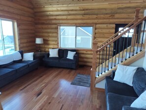 A log cabin interior with a wooden staircase, a dark-colored sofa set, and a large window. - Cozy Cottage 5-bedrooms, Ideal Beach Resort access, view of Bear Lake, sleeps 16 (Garden City)