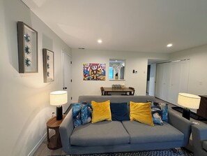 A modern living room with a grey sofa, two lamps, a coffee table, and a colorful artwork on the wall. - Charming Cabin | 3BR, Epic Game Room and Fireplace (Hermantown)
