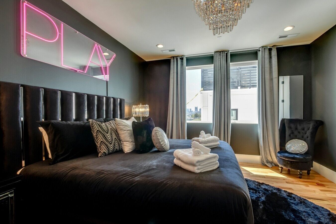 A modern bedroom with a large bed, a neon sign, a chandelier, and a view of the city.