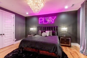Um quarto com uma cama, mesas de cabeceira, um candelabro e um letreiro de neon com a palavra 'PLAY' na parede.