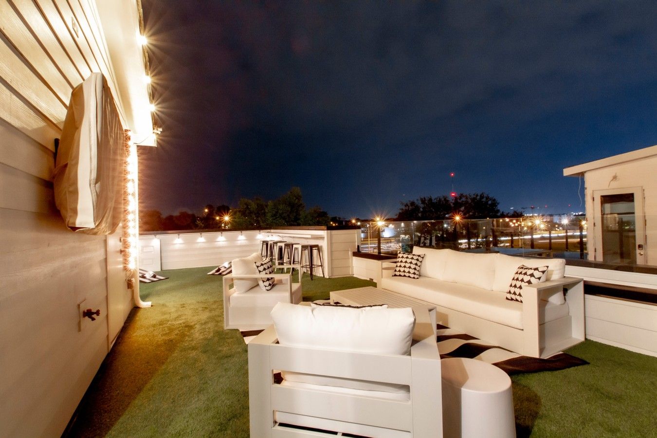 An outdoor seating area with white furniture and artificial grass, overlooking a cityscape at night.
