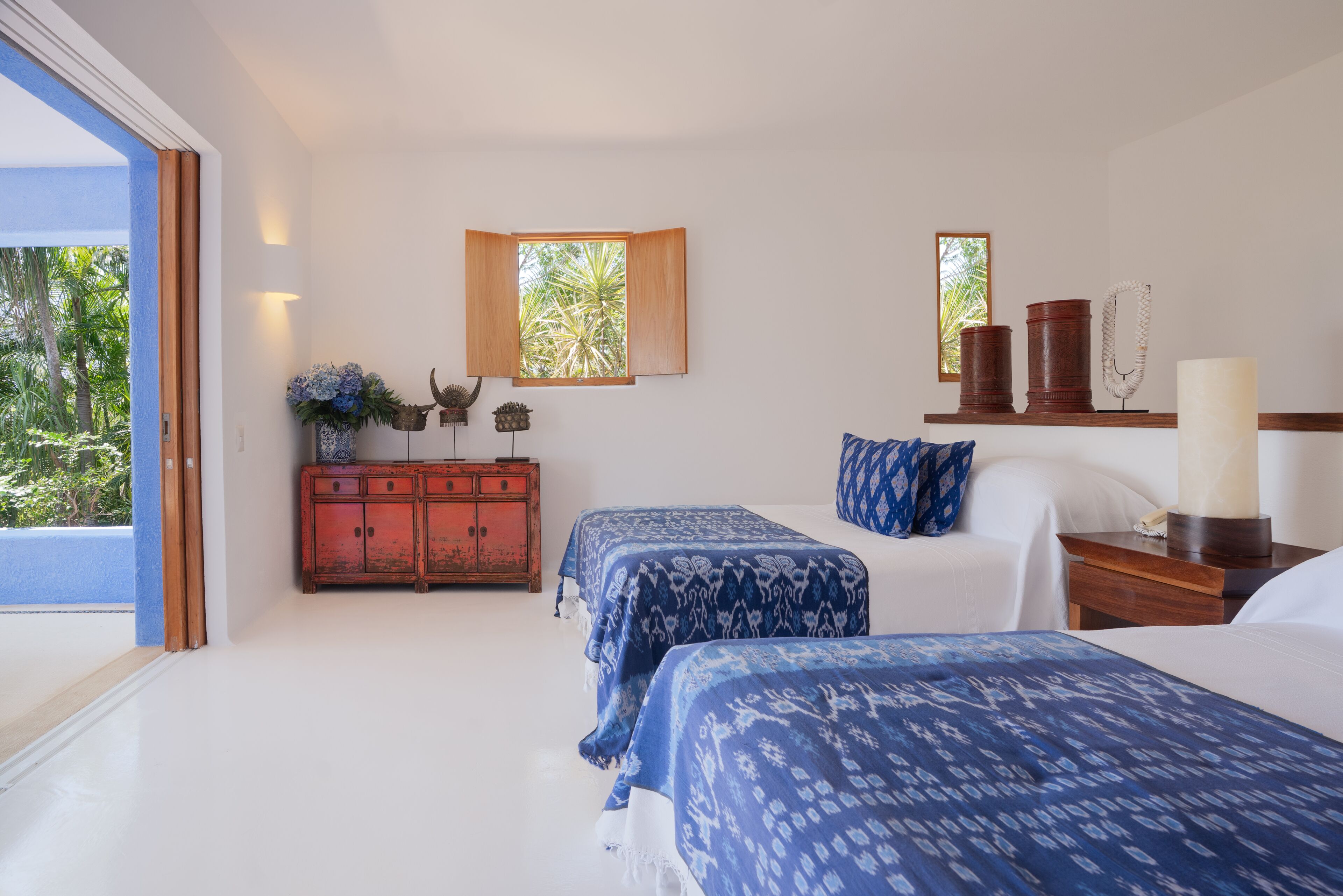 Un dormitorio con dos camas, un armario de madera, una ventana con vistas a palmeras y una mesita de noche con una lámpara.