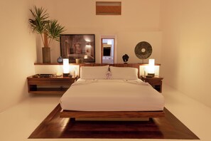Quarto com cama de madeira, mesas de cabeceira com luminárias, um vaso de planta, um quadro e um prato decorativo na parede.