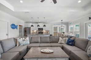 A modern living room with a sectional sofa, a coffee table, pendant lights, and a kitchen in the background. - Caribbean Surf | Private Pool | Beach Access | PCB | Near Beach (Panama City Beach)