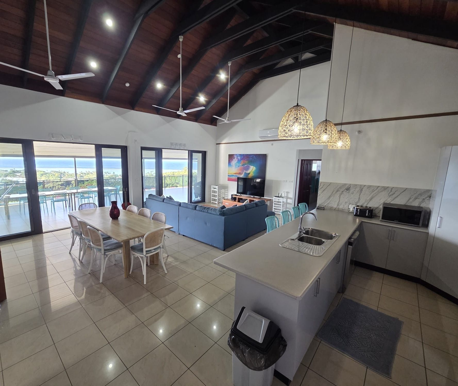 A modern kitchen with a central island, a dining area with a table and chairs, and large windows offering a view of the outdoors.