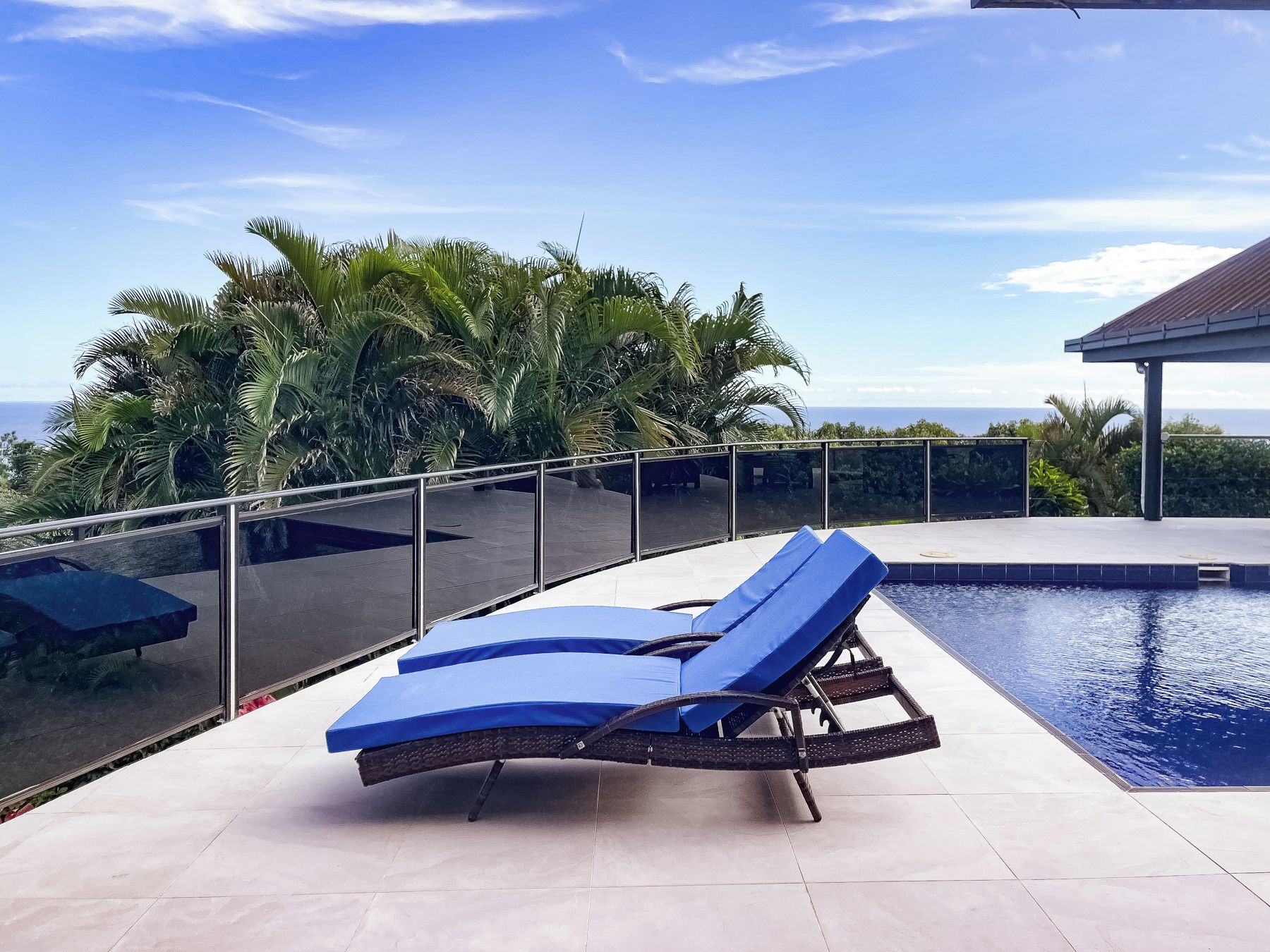 Un espace aménagé au bord de la piscine, avec un fauteuil de relaxation et une balustrade en verre donnant sur une étendue d’eau et des palmiers.