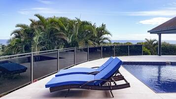 Un espace aménagé au bord de la piscine, avec un fauteuil de relaxation et une balustrade en verre donnant sur une étendue d’eau et des palmiers.