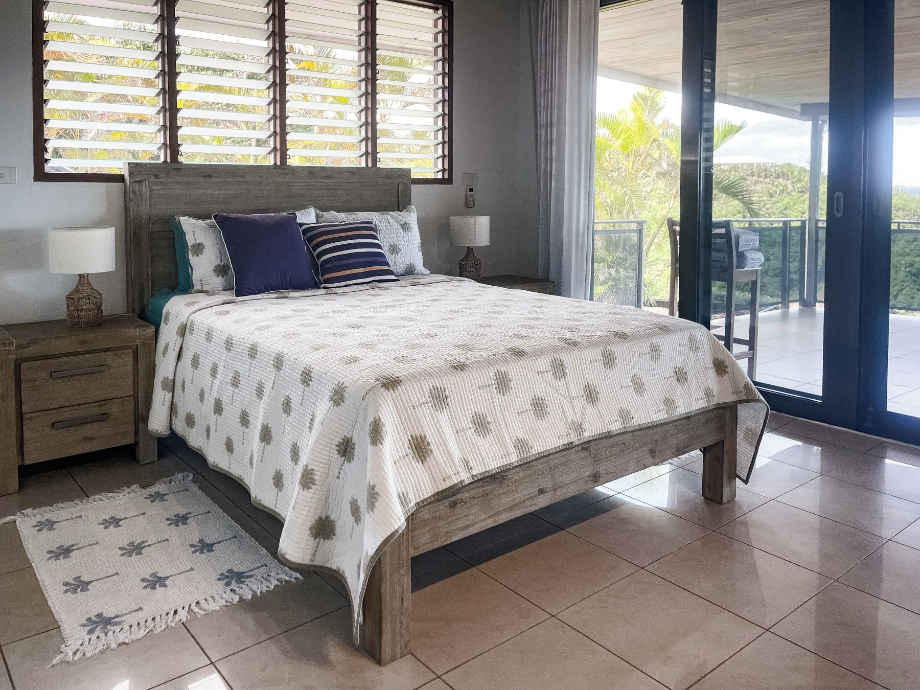 A bedroom with a bed, bedside tables, a nightstand, a window with blinds, a balcony, and a view of greenery.