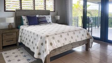 A bedroom with a bed, bedside tables, a nightstand, a window with blinds, a balcony, and a view of greenery.