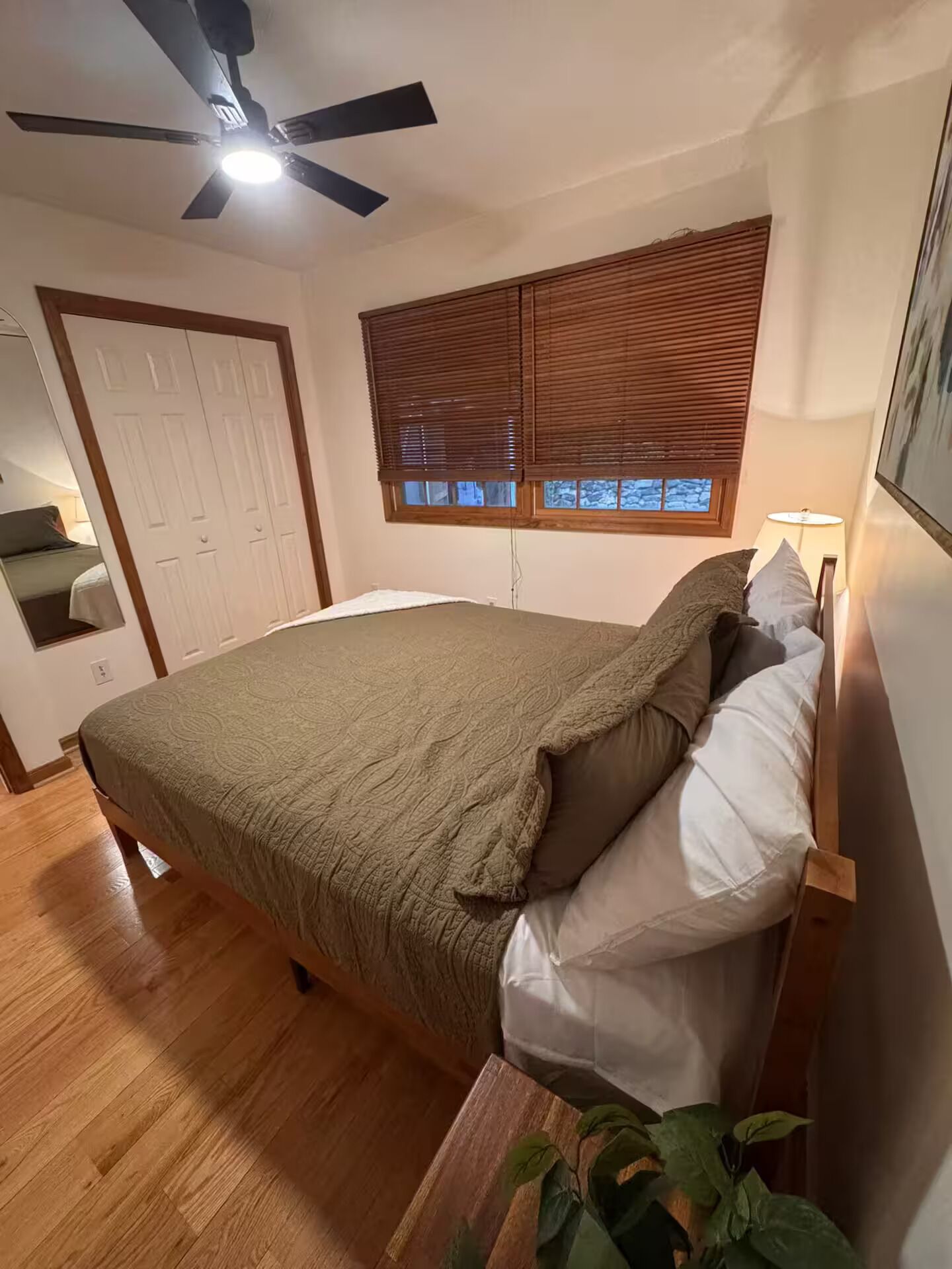 A bedroom with a bed, wooden floor, ceiling fan, window blinds, and a bedside lamp.