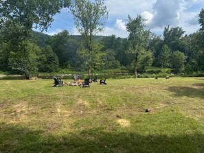 An outdoor seating area with chairs and tables, surrounded by trees and a body of water. - Modern Riverfront Retreat - Hot Tub & Fire Pit - Minutes from Berkley Springs (Great Cacapon)