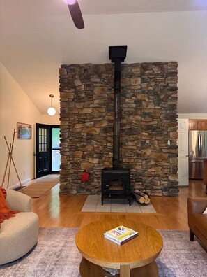 A cozy living room with a stone fireplace, a round wooden coffee table, and a bookshelf. - Modern Riverfront Retreat - Hot Tub & Fire Pit - Minutes from Berkley Springs (Great Cacapon)