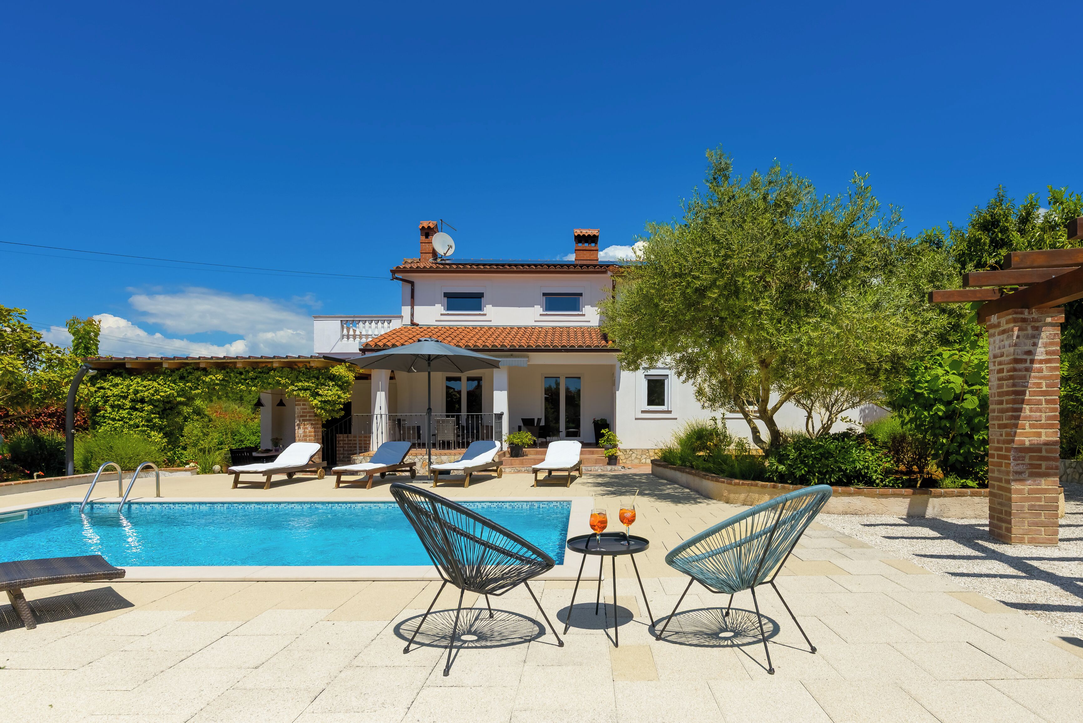 Una piscina con sillas de descanso y una mesa, una casa con techo de tejas y un cielo azul despejado.