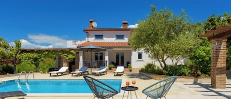Una piscina con sillas de descanso y una mesa, una casa con techo de tejas y un cielo azul despejado.