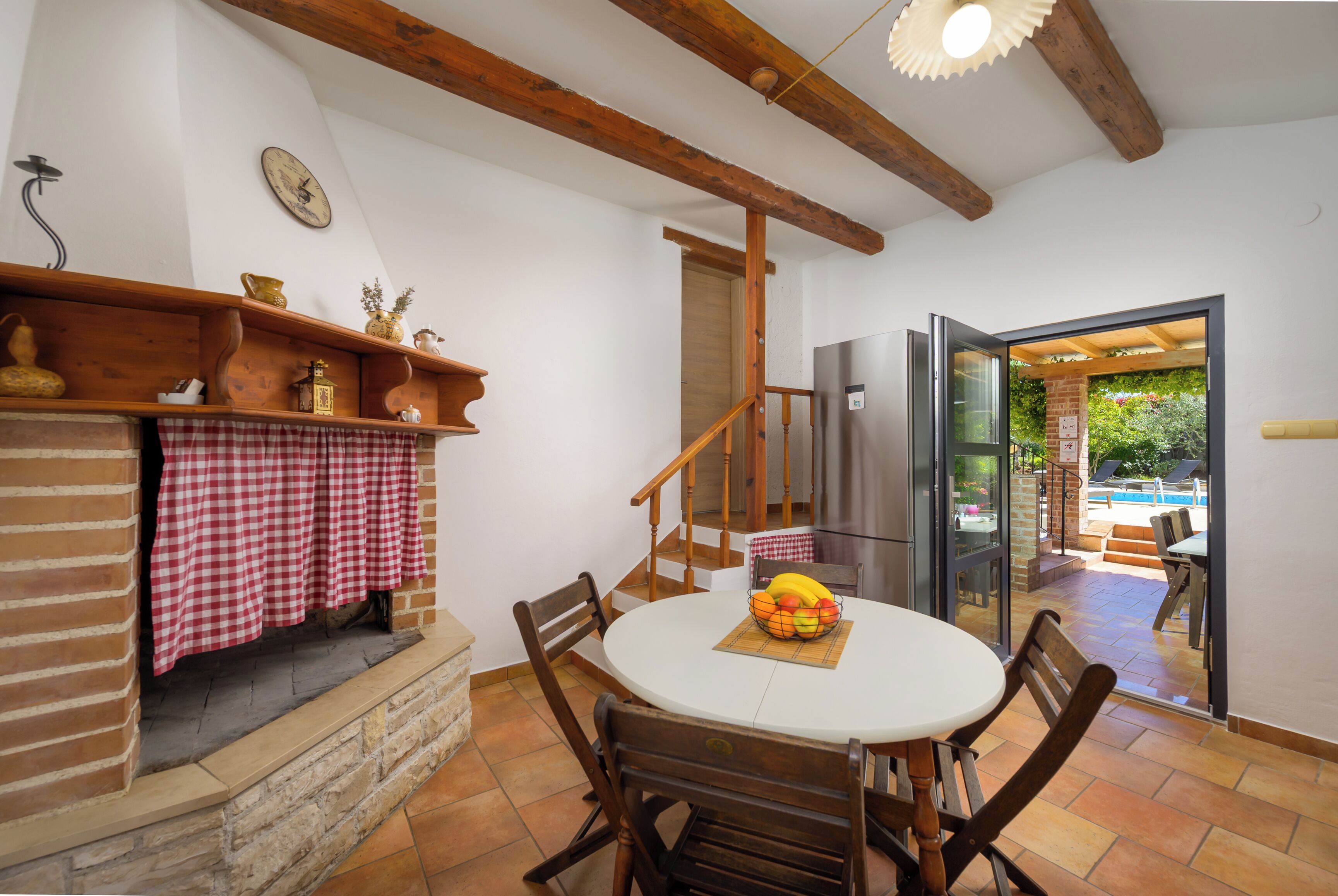 A dining area with a round table, chairs, a checkered tablecloth, a fruit basket, and a view of an outdoor patio with a pool.