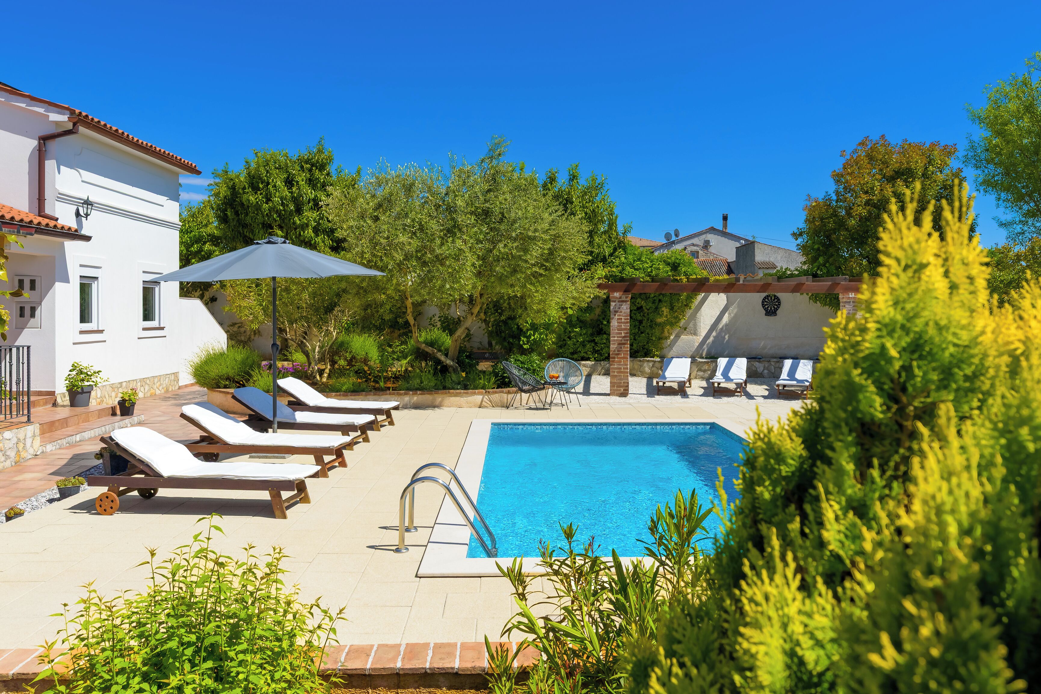 A swimming pool with lounge chairs and a pergola, surrounded by greenery and a white building.