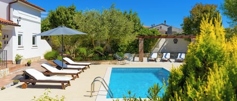 A swimming pool with lounge chairs and a pergola, surrounded by greenery and a white building.
