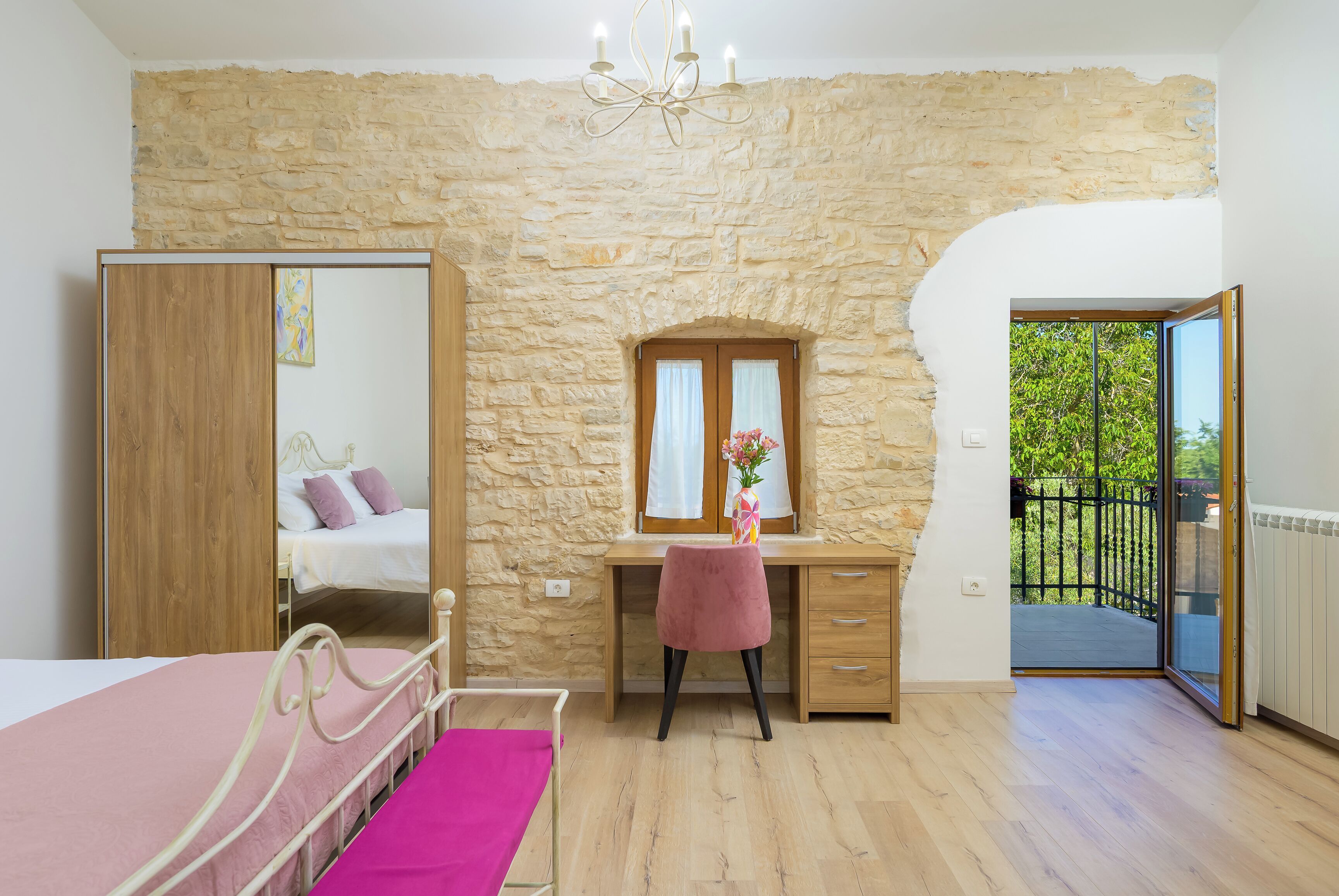 A bedroom with a stone wall, a bed, a desk with a chair, a mirror, and a balcony.