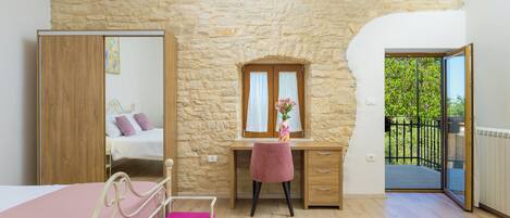 A bedroom with a stone wall, a bed, a desk with a chair, a mirror, and a balcony.
